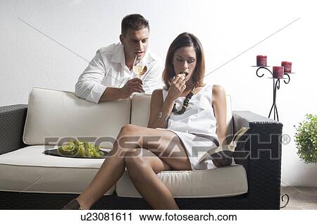 Stock Image - Young man leaning on couch with white wine and young woman reading book and eating grapes. Fotosearch