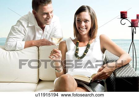 Picture - Young man leaning on couch with young woman with book and white wine. Fotosearch