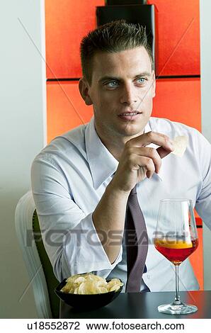 Stock Image - Young man sitting with a glass of orange wine and potato chips. Fotosearch