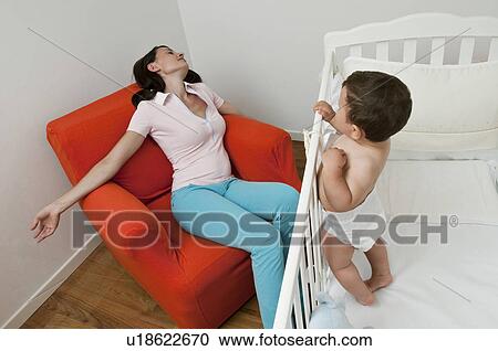 Stock Image - Young woman asleep on arm chair and baby boy in crib. Fotosearch
