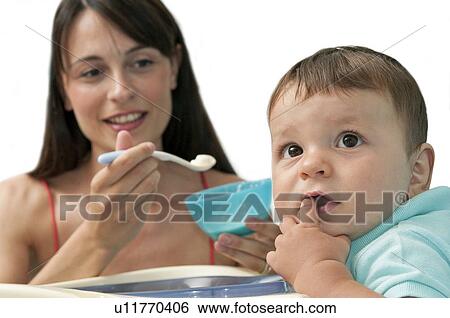 Stock Photograph - Young woman feeding baby boy in high chair. Fotosearch