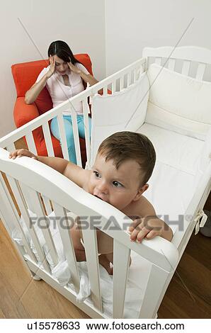 Stock Image - Young woman on arm chair holding her head and baby boy in crib. Fotosearch