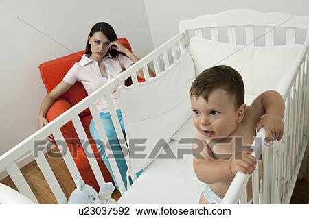 Young woman on arm chair holding her head and baby boy in crib View Large Photo Image Stock Image - Young woman on arm chair holding her head and baby boy in crib. Fotosearch