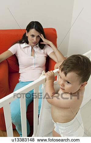 Stock Photo - Young woman on arm chair holding her head and baby boy in crib. Fotosearch