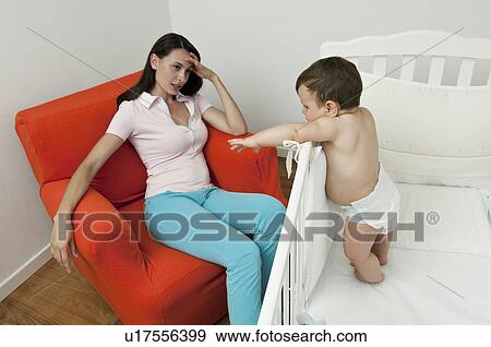 Stock Photo - Young woman on arm chair holding her head and baby boy in crib. Fotosearch