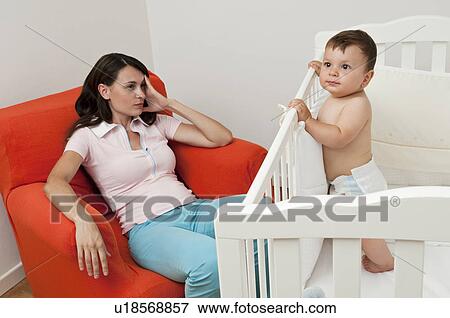 Stock Photo - Young woman on arm chair holding her head and baby boy in crib. Fotosearch