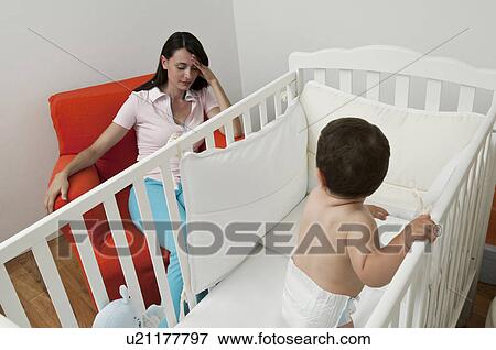 Stock Photo - Young woman on arm chair holding her head and baby boy in crib. Fotosearch