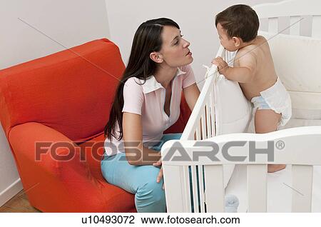 Stock Image - Young woman on arm chair looking at baby boy in crib. Fotosearch