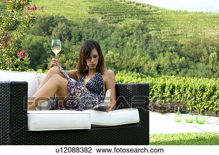 Stock Image - Young woman sitting on couch in vineyard with white wine reading a book. Fotosearch
