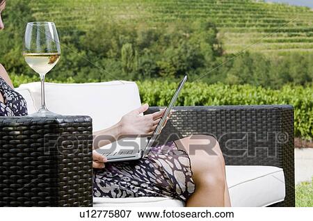 Stock Photo - Young woman sitting on couch in vineyard with white wine and laptop. Fotosearch