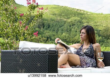 Stock Photo - Young woman sitting on couch in vineyard with white wine reading a book. Fotosearch