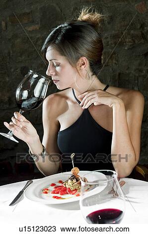 Stock Image - Young woman with plate of food sniffing her wine at restaurant. Fotosearch