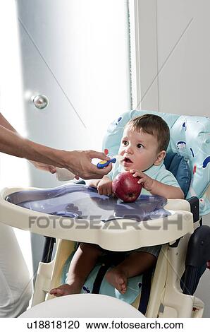 Stock Image - Young woman's hand feeding baby boy in high chair holding an apple. Fotosearch