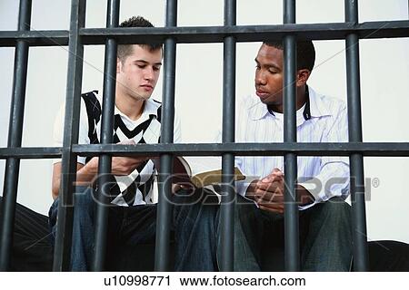 Stock Image - a young man reads the bible to another young man in jail. Fotosearch