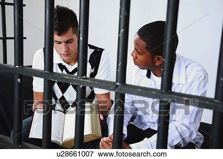 Stock Photo - a young man reads the bible to another young man in jail. Fotosearch