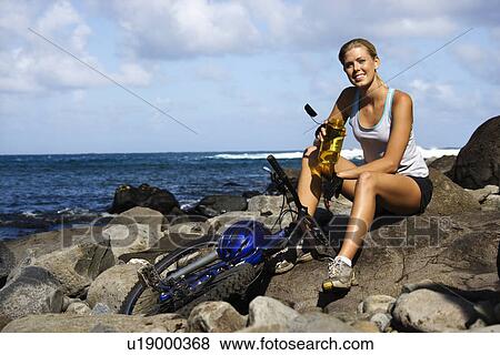 Stock Photo - Attractive Young Woman Sitting With Bicycle on Rocky Beach. Fotosearch