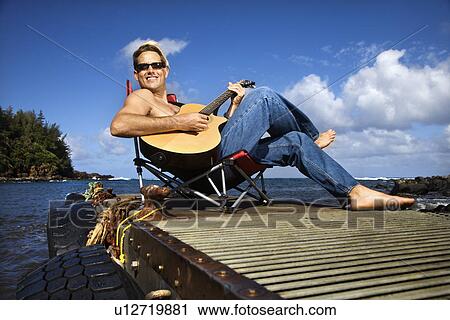 Stock Image - Young Man Sitting Lakeside and Playing Guitar. Fotosearch