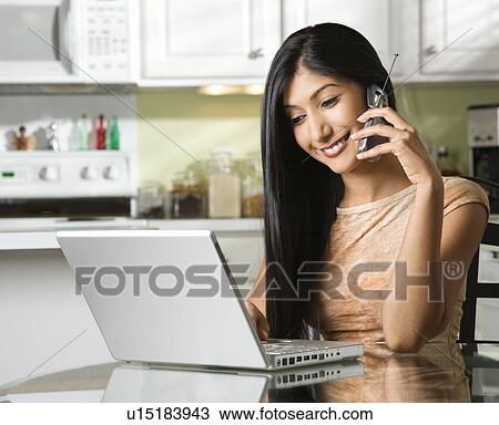 Stock Image - Young Woman Using Laptop and Talking on Cellphone. Fotosearch
