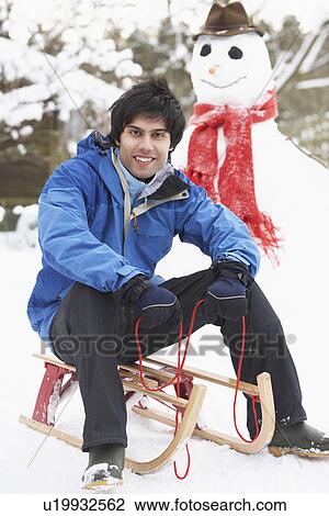 Stock Image - Teenage Boy With Sledge Next To Snowman. Fotosearch