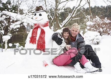 Stock Image - Teenage Couple In Winter Landscape Next To Snowman With Flask And Hot Drink. Fotosearch