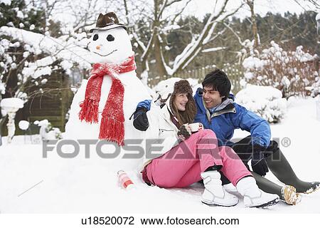 Stock Image - Teenage Couple In Winter Landscape Next To Snowman With Flask And Hot Drink. Fotosearch