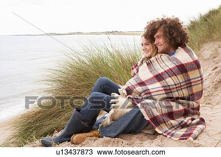 Stock Image - Teenage Couple Sitting In Sand Dunes Wrapped In Blanket. Fotosearch