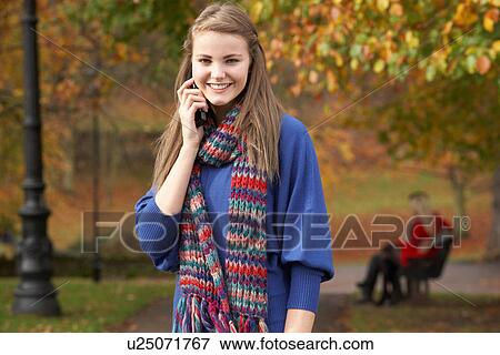Stock Photo - Teenage Girl On Mobile Phone In Autumn Park With Couple On Bench In Background. Fotosearch