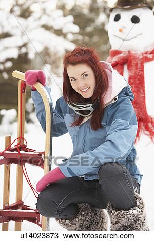Stock Image - Teenage Girl With Sledge Next To Snowman. Fotosearch