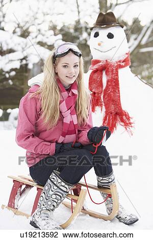 Stock Image - Teenage Girl With Sledge Next To Snowman. Fotosearch