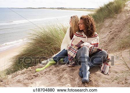 Stock Image - Two Teenage Girls Sitting In Sand Dunes Wrapped In Blanket. Fotosearch