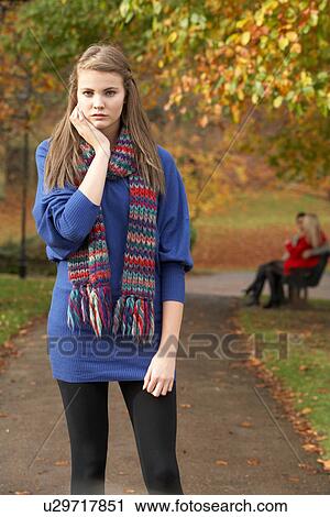Stock Image - Unhappy Teenage Girl Standing In Autumn Park With Couple On Bench In Background. Fotosearch