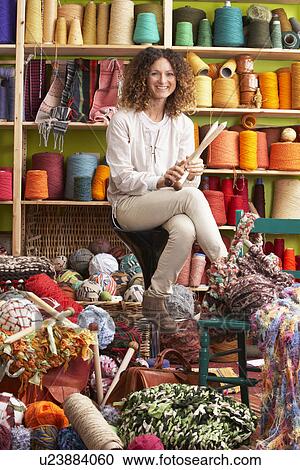 Stock Image - Woman Sitting On Stool Holding Knitting Needles In Front Of Yarn Display. Fotosearch