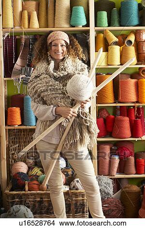 Picture - Woman Wearing Knitted Scarf Standing In Front Of Yarn Display Holding Giant Needles And Ball Of Wool. Fotosearch