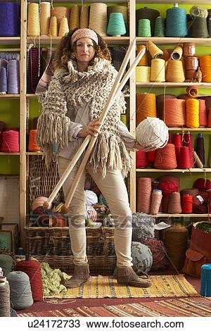 Stock Image - Woman Wearing Knitted Scarf Standing In Front Of Yarn Display Holding Giant Needles. Fotosearch