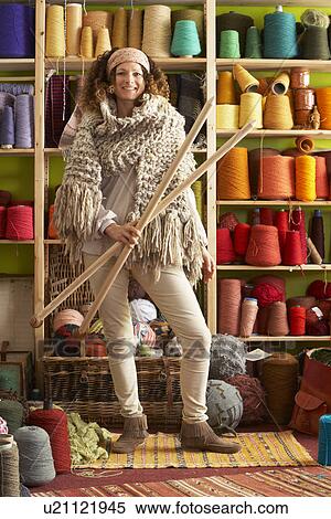 Stock Photography - Woman Wearing Knitted Scarf Standing In Front Of Yarn Display Holding Giant Needles. Fotosearch