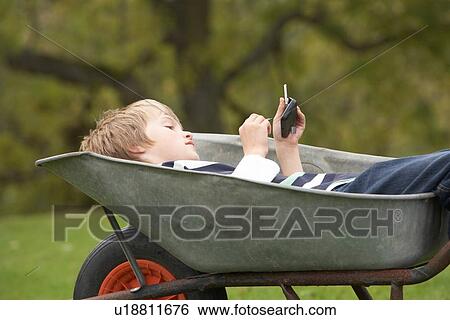 Stock Photograph - Young Boy Laying Wheelbarrow Using Smart Mobile Phone. Fotosearch