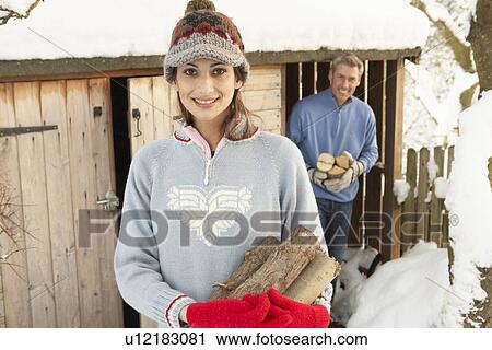 Stock Image - Young Couple Collecting Logs From Wooden Store In Snow. Fotosearch