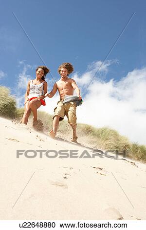 Stock Image - Young Couple Running Down Sand Dune On Beach. Fotosearch