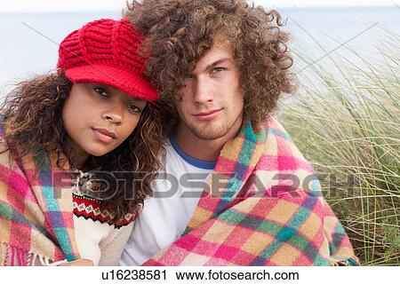 Stock Image - Young Couple Sitting In Sand Dunes Wrapped In Blanket. Fotosearch