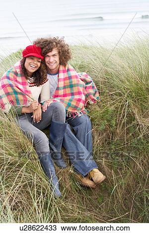 Stock Image - Young Couple Sitting In Sand Dunes Wrapped In Blanket. Fotosearch