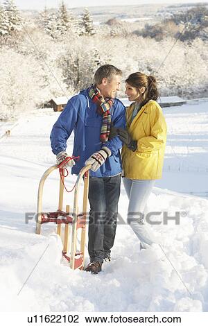 Stock Image - Young Couple Standing In Snowy Landscape Holding Sledge. Fotosearch