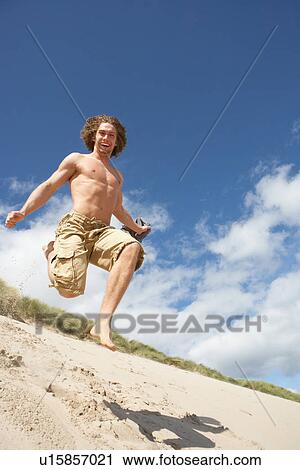 Stock Image - Young Man Running Down Sand Dune On Beach. Fotosearch