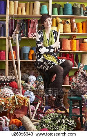Stock Image - Young Woman Sitting On Stool Holding Knitting Needles In Front Of Yarn Display. Fotosearch