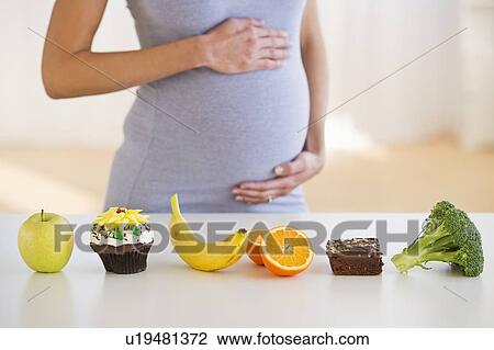 Stock Image - Pregnant woman standing behind a row of food. Fotosearch