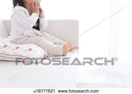 Stock Image - Baby girl sitting on cushion on sofa. Fotosearch