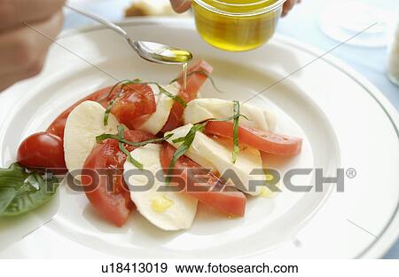 Stock Photo - Hand pouring olive oil on tomatoes, mozzarella and basil salad. Fotosearch