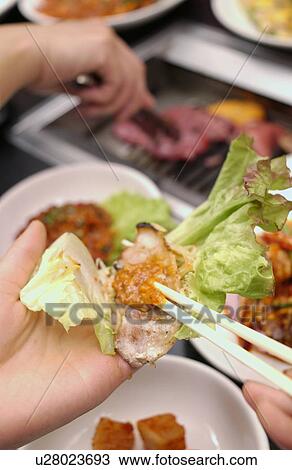 Stock Image - Hands wrapping slice of meat with lettuce, close up. Fotosearch