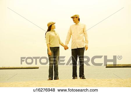 Stock Photo - Mature couple in flat cap standing by sea, hand in hand. Fotosearch
