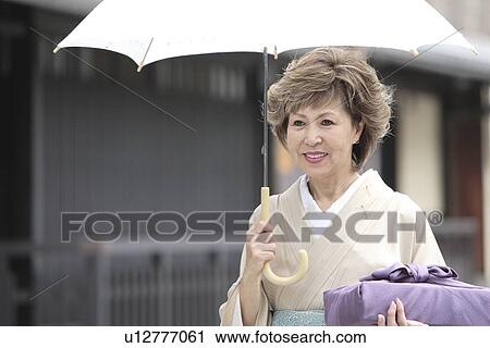 Stock Image - Mature woman in Kimono holding umbrella and gift, Kyoto city, Kyoto prefecture, Japan. Fotosearch