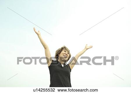 Stock Image - Mature woman raising arms under sky, smiling. Fotosearch